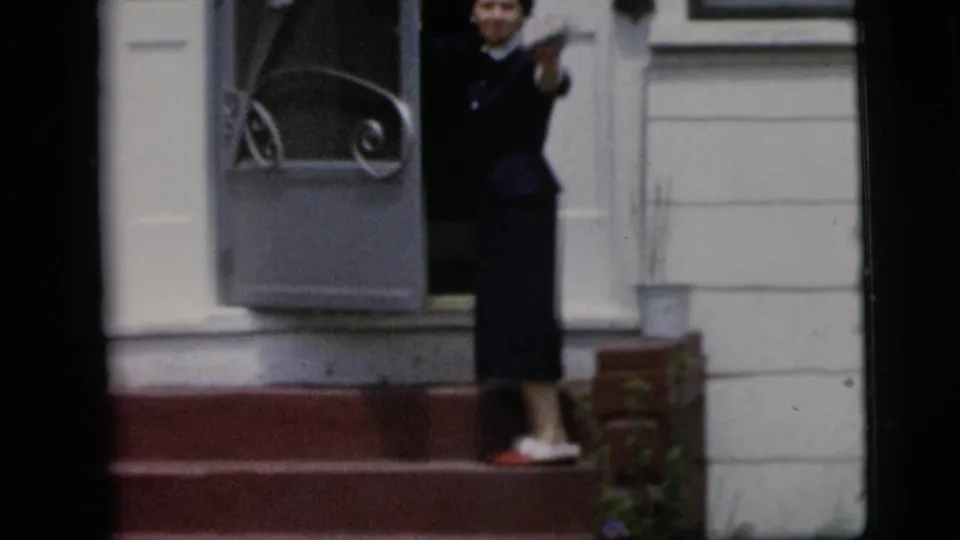 The woman turns back at the doorway and waves toward the camera in a 1958 New York City residential home-movie scene.