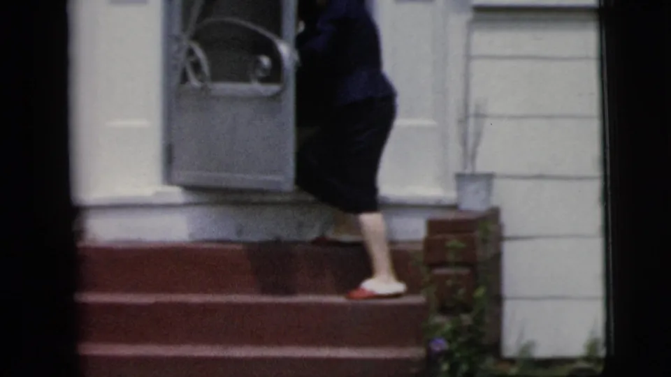 The woman steps through the gray storm door into the white house, closing the brief 1958 New York City home entrance moment.