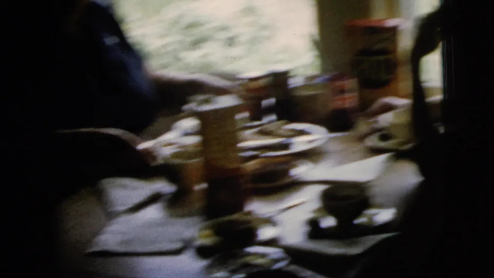 A dim 1958 New York City interior shows hands around a dining table with cups, plates, and a cracker box near the window.