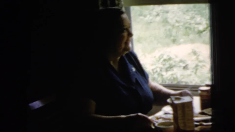 A woman sits beside a sunlit window in a New York City home, with plates, cups, and a cracker box on the table.