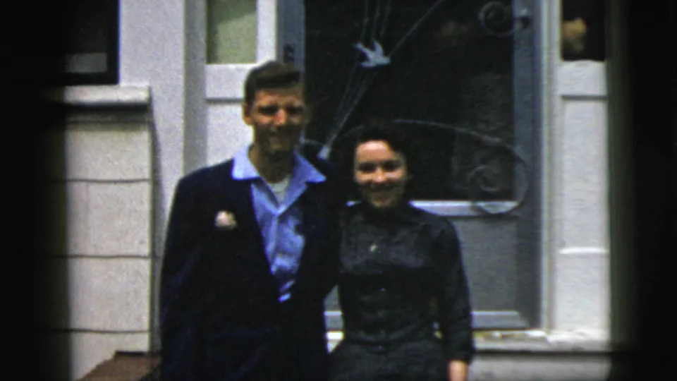 The couple smiles toward the camera outside a New York City building, with doorway trim and decorative glass behind them.