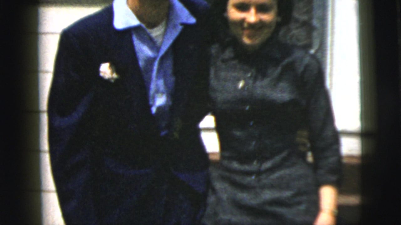 The couple walks together outside a New York City house, with the man wearing a boutonniere and the woman in a dark dress.