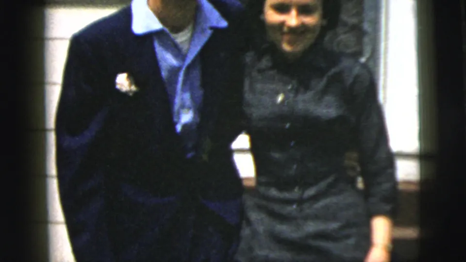 The couple walks together outside a New York City house, with the man wearing a boutonniere and the woman in a dark dress.