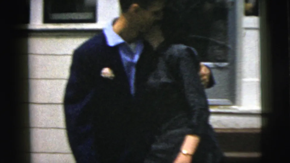 The couple leans in for a kiss outside a New York City residence, framed in soft 8mm home-movie focus.