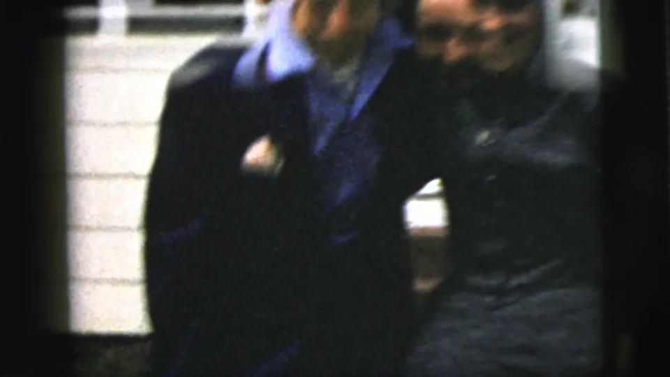 The couple appears to laugh and lean together in front of a residential exterior, with motion blur and handheld 8mm framing.
