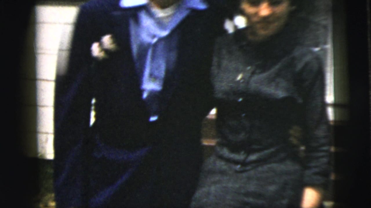 Smiling couple close together, with a dark suit, blue collar, and dark dress visible.