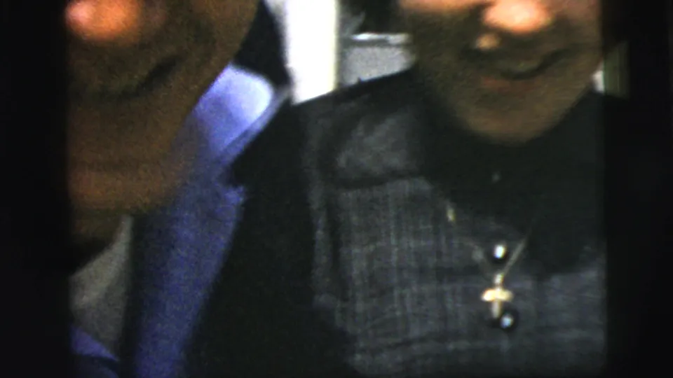 A close 1958 New York City home-movie frame shows the woman smiling in a dark dress while the suited man is partly cropped beside her.