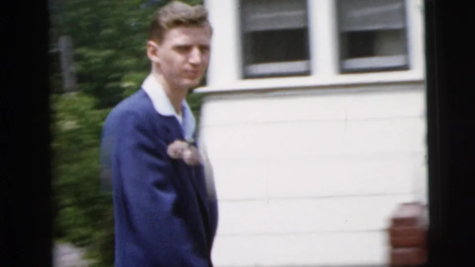 Teenage boy in a blue suit and white shirt, with a lapel corsage, walking outside a New York City house in 1958 footage.