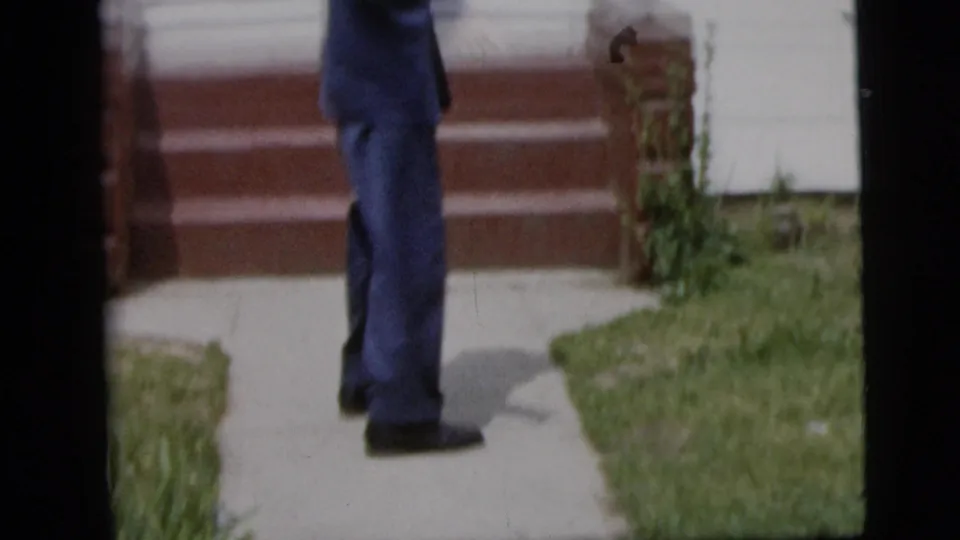 Lower portion of the teenage boy in a blue suit near a sidewalk, lawn, and red porch steps in 1958 home-movie footage.