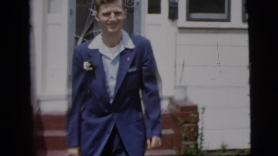 Teenage boy in a blue suit and lapel corsage smiles near the front porch of a white house in 1958 New York City footage.
