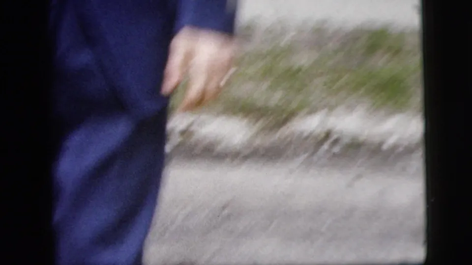 The man in the blue suit passes close to the handheld camera, with sidewalk and curbside greenery visible behind him.