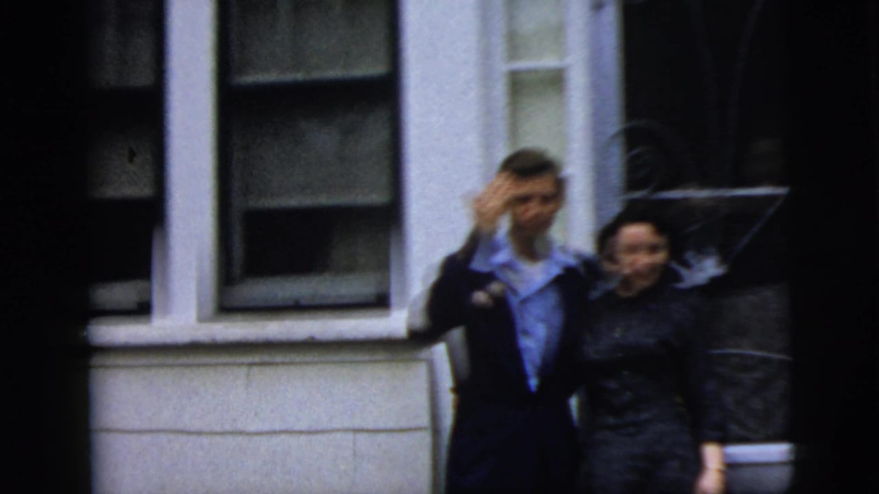 Young couple in formal clothing appears outside a New York City residence, with the man raising a hand toward the camera.
