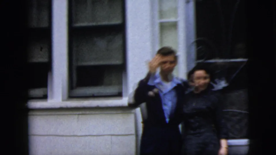Young couple in formal clothing appears outside a New York City residence, with the man raising a hand toward the camera.