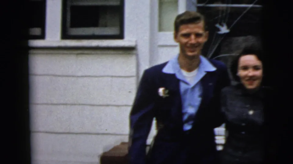 A smiling formally dressed couple approaches the camera outside a New York City residence, with porch details behind them.