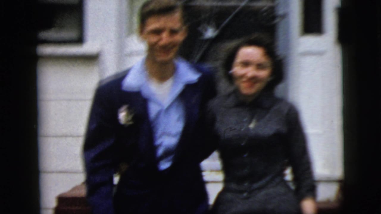 In 1958 New York City home-movie footage, a young man in a dark suit and a woman in a dark dress smile near a house exterior.