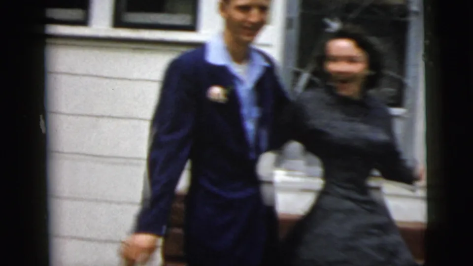The dressed-up couple dances hand in hand outside a New York City residence, captured with soft focus and handheld movement.