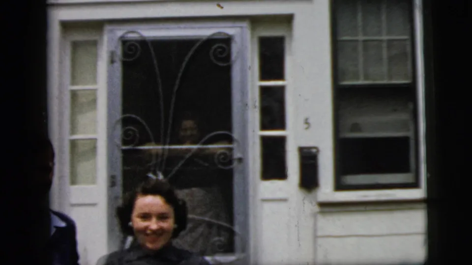 The woman smiles near the front entrance of a white New York City house, with the decorative screen door and number visible.