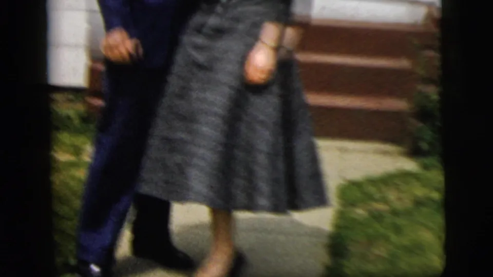 The formally dressed couple stands together outside the home, with porch steps and a small lawn visible in the frame.
