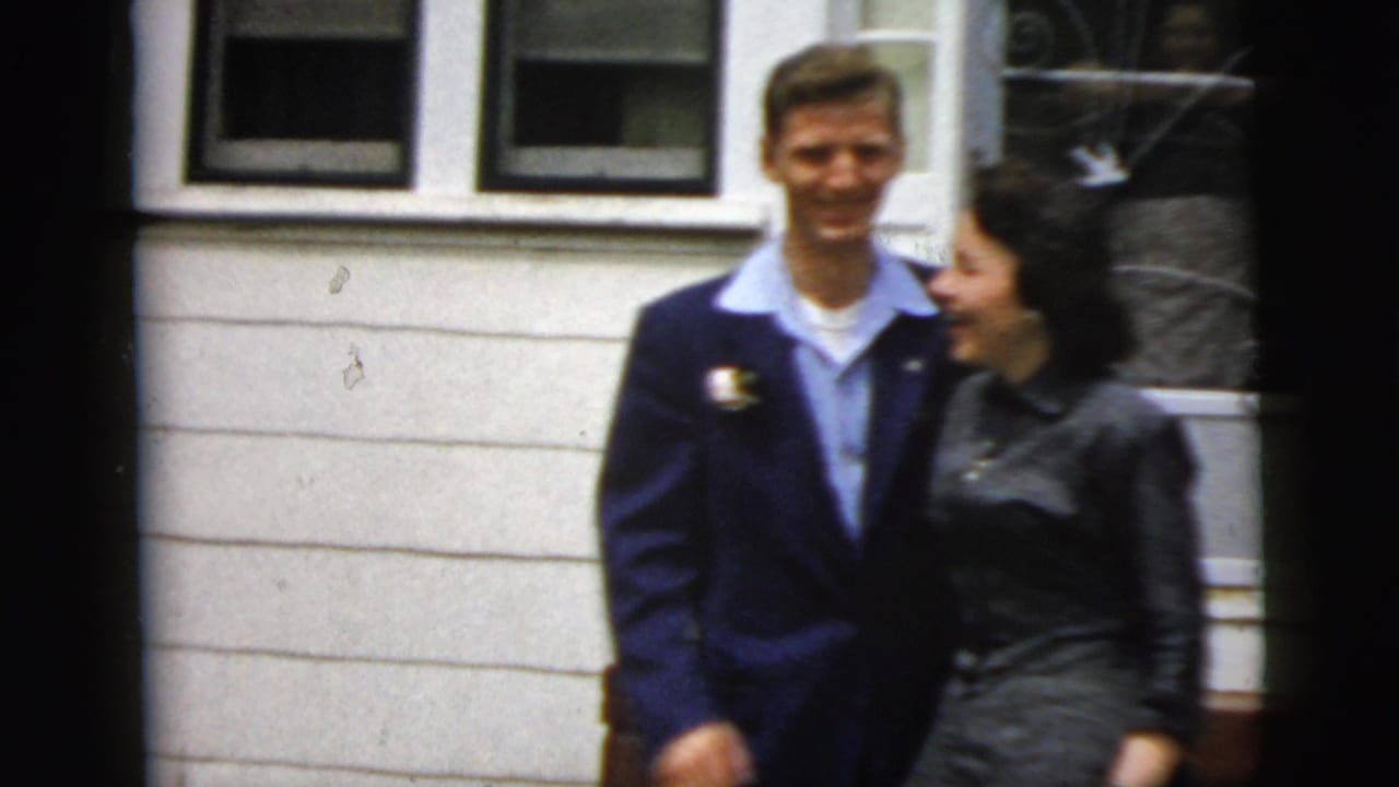 Young man in a dark suit stands with a woman outside a white-sided house.