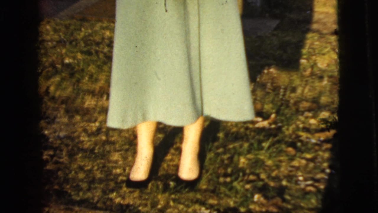 Lower half of a woman in a light coat and dark shoes standing on grass.