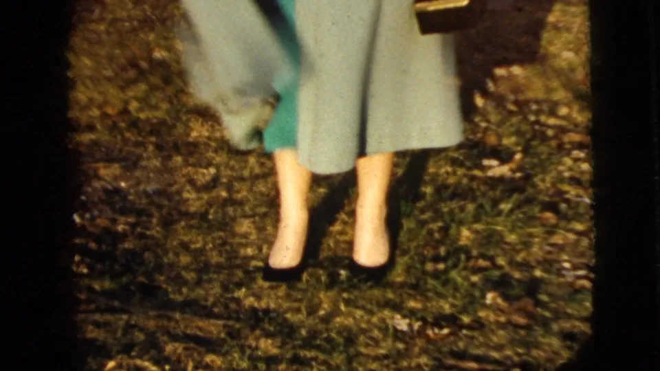 The handheld camera remains low on the woman's outfit, showing the light coat, green dress, dark shoes, purse edge, and uneven grassy ground.