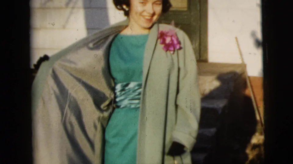 A smiling woman in a green dress, light coat, and pink corsage stands outside near house steps, holding or wearing a small dark purse.