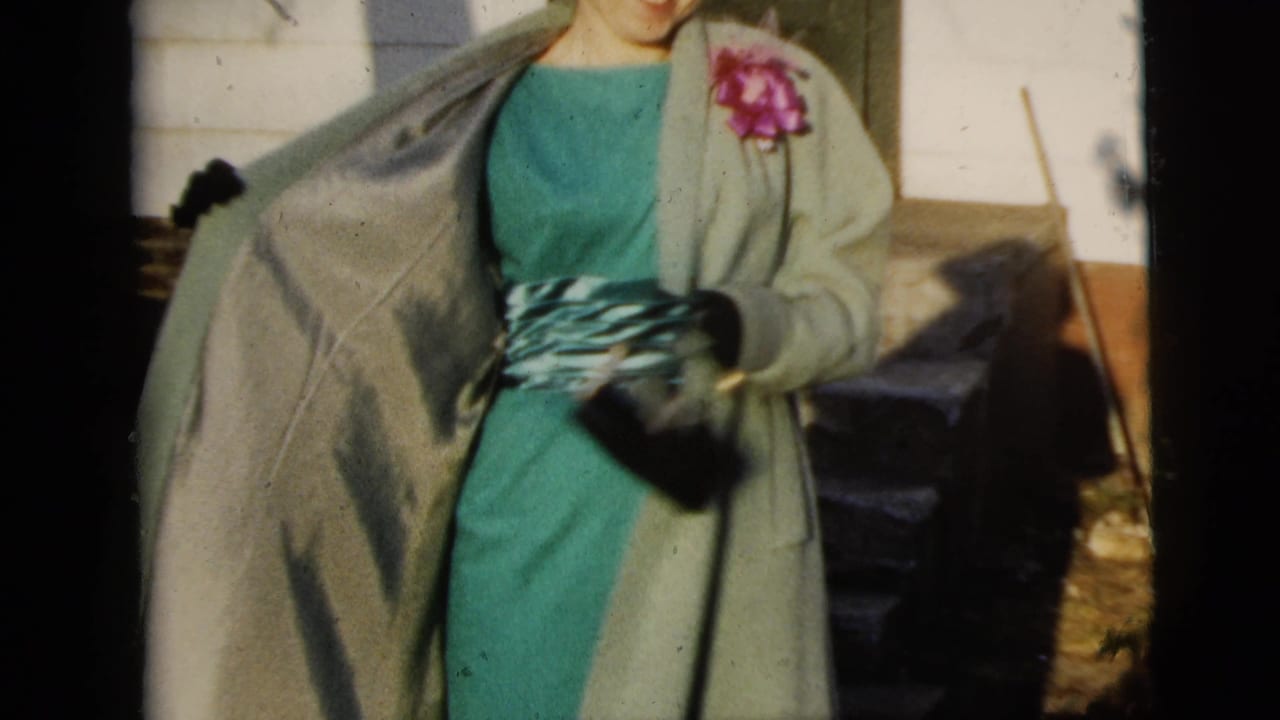 Woman in a teal dress and light coat poses outdoors with gloves and a purse.