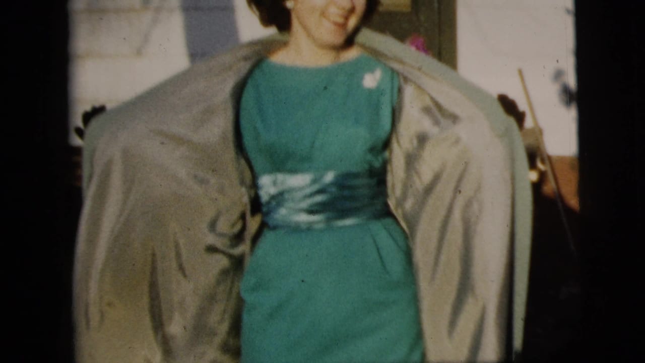 Smiling woman opens a light coat to reveal a teal dress and sash.