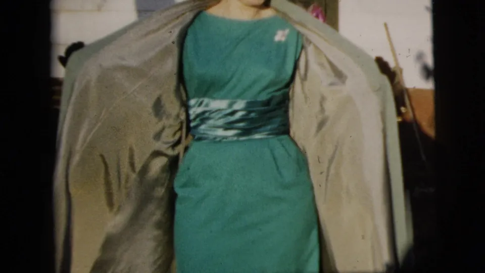 The home-movie frame crops closely on the woman's teal dress, patterned sash, and open light coat.