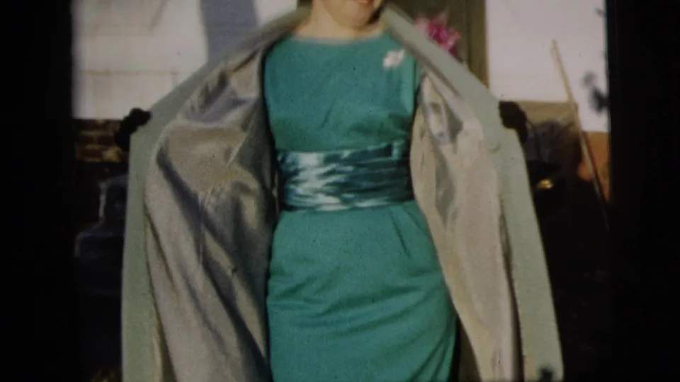 A woman in a teal dress and light coat stands in bright outdoor light near residential steps in New York City footage.