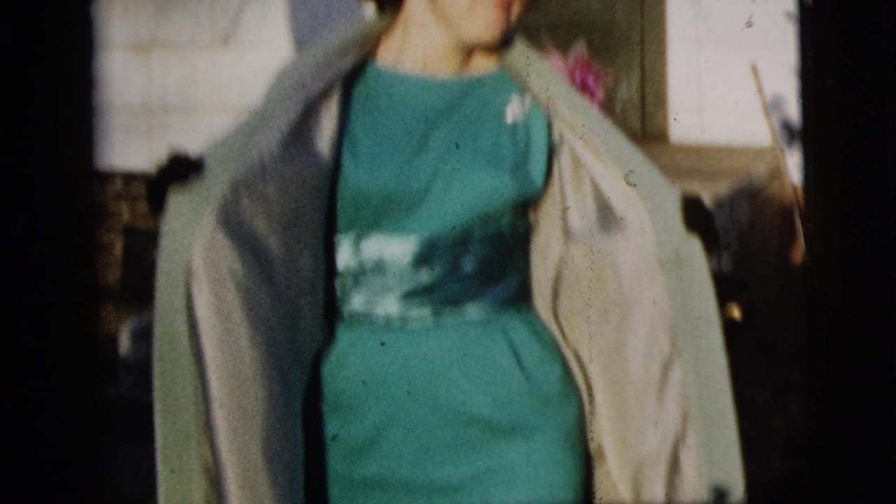 In 1958 New York City home-movie footage, a woman in a teal dress and light coat moves toward the camera outside a clapboard house.