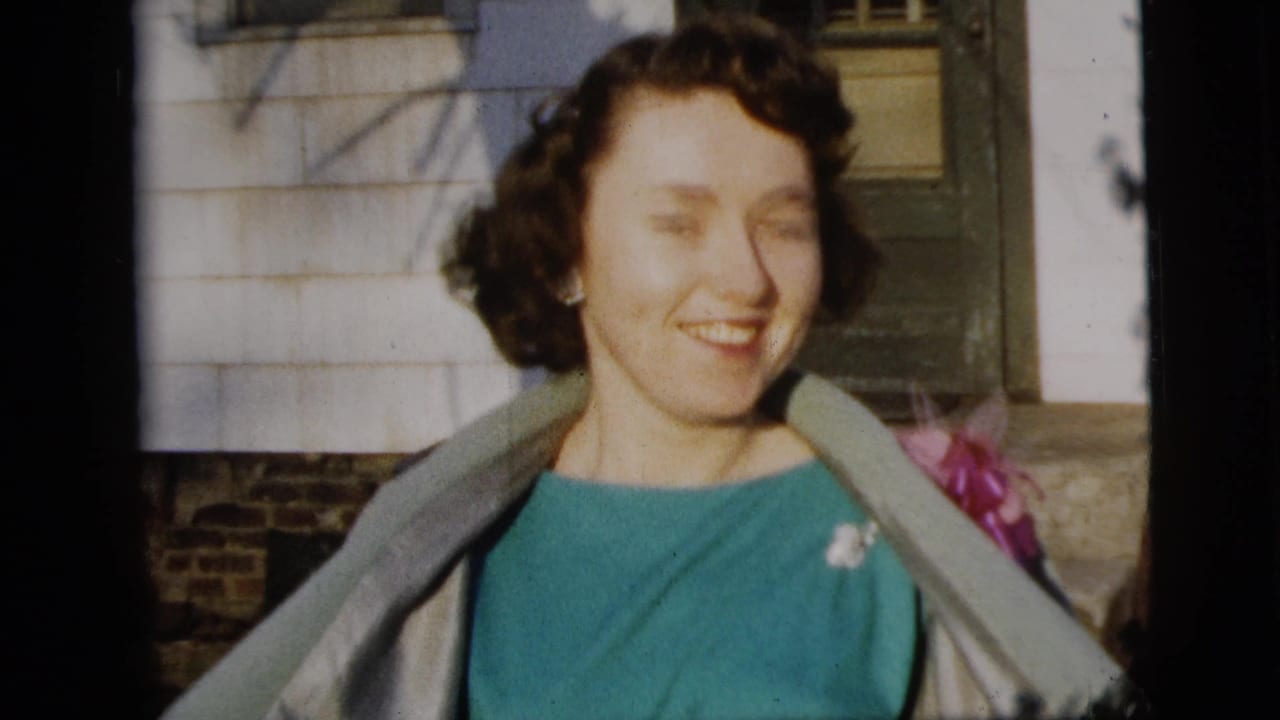 A smiling woman in a teal dress, light coat, and pink corsage stands outside a New York City residence in 1958 home-movie footage.