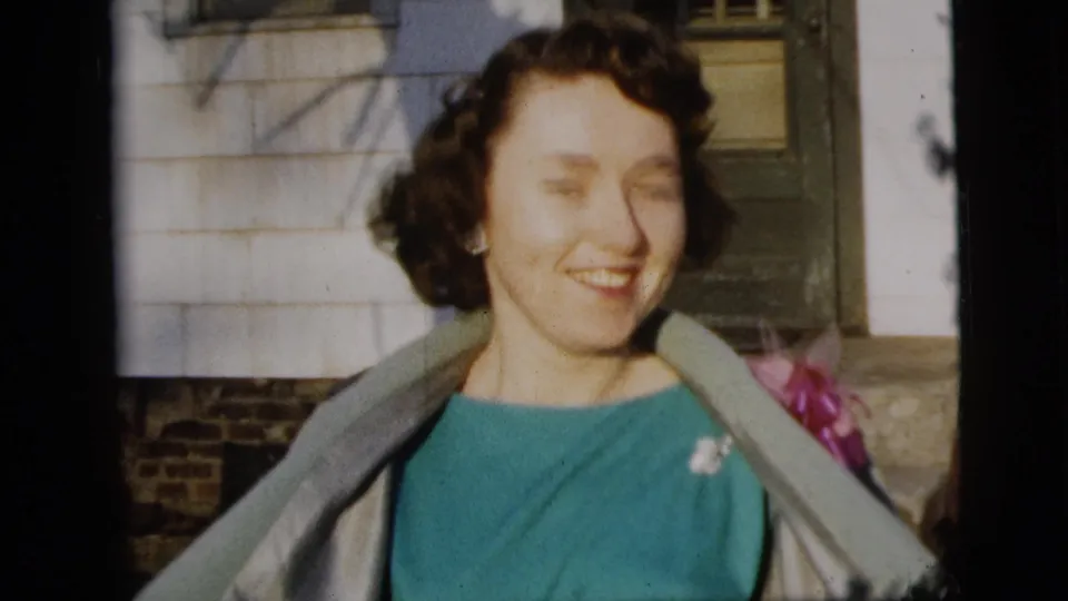 A smiling woman in a teal dress, light coat, and pink corsage stands outside a New York City residence in 1958 home-movie footage.