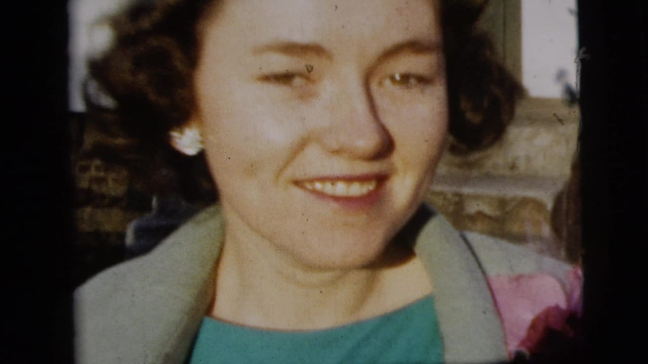 Close-up of a smiling woman with curled hair, pearl earrings, teal dress, and light coat in 1958 New York City home-movie footage.