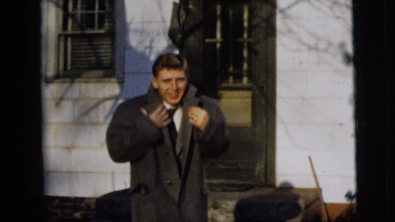 A man in a dark overcoat adjusts his tie or lapels outside a clapboard house in 1958 New York City home-movie footage.