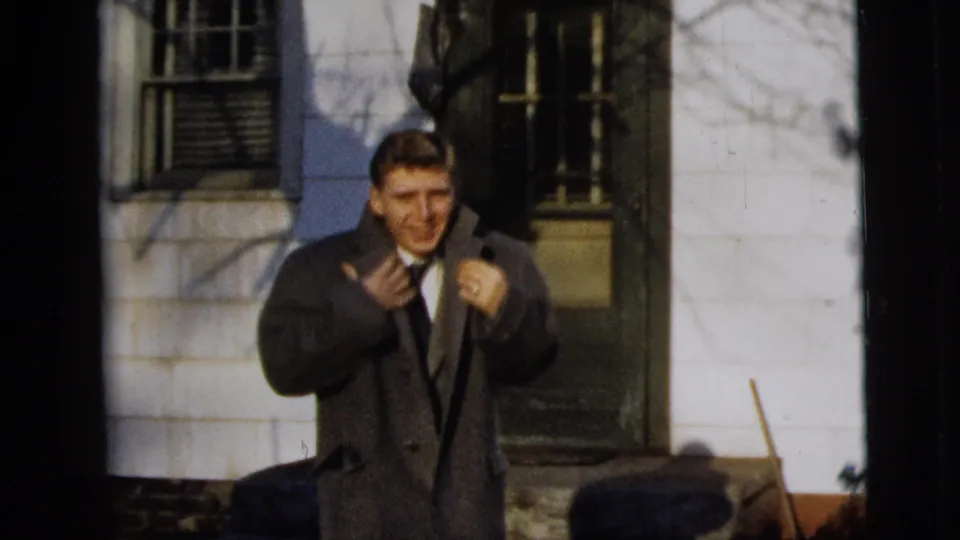 A man in a dark overcoat adjusts his tie or lapels outside a clapboard house in 1958 New York City home-movie footage.