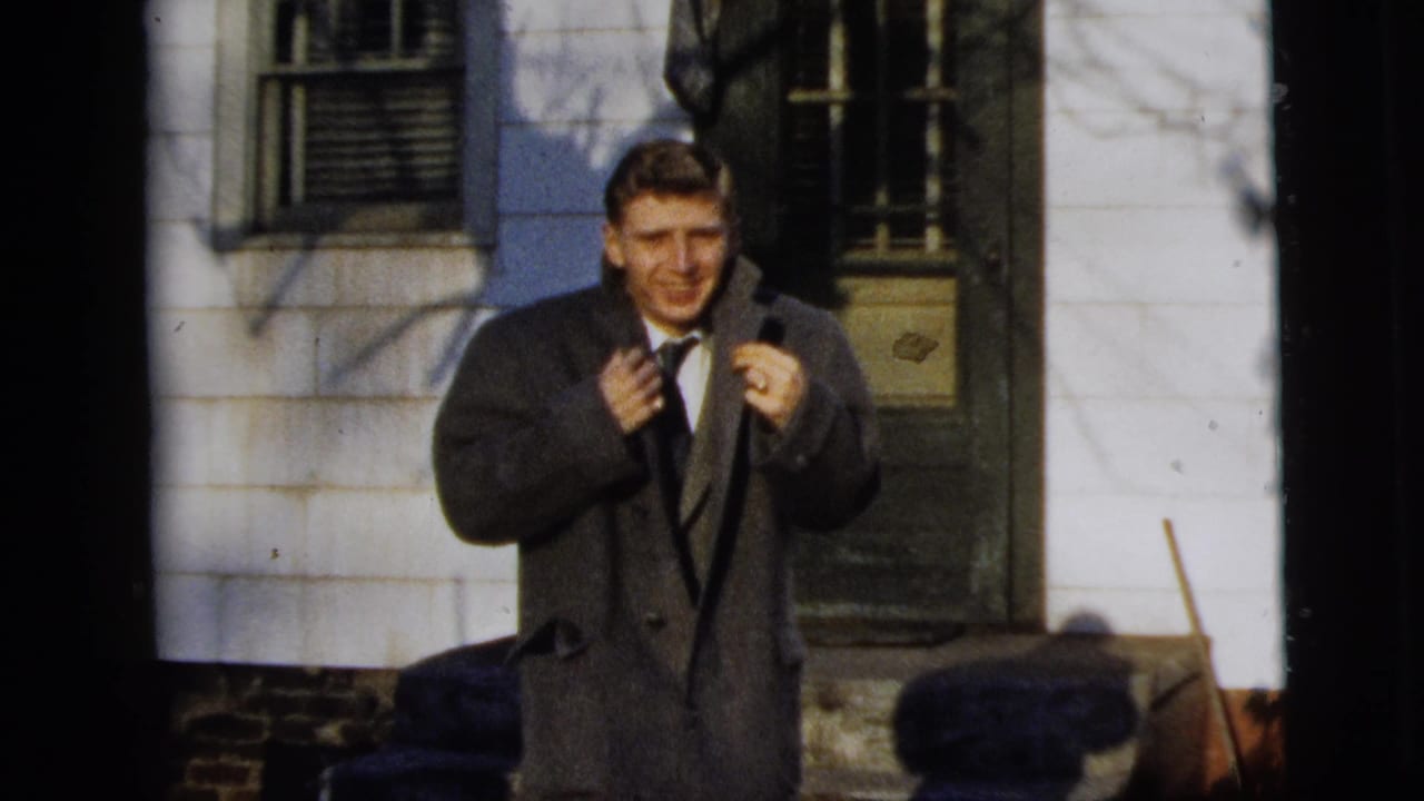 In 1958 New York City home-movie footage, a man in a dark overcoat and tie smiles toward the camera outside a house.