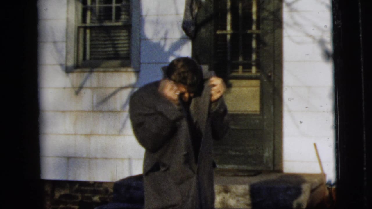 Man lowers his head while adjusting his coat collar outside a house.