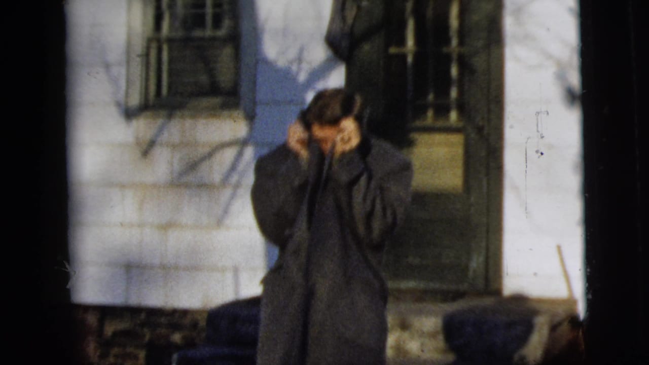 The man raises both hands near his face and collar, partly hiding his expression in the 1958 exterior home-movie scene.