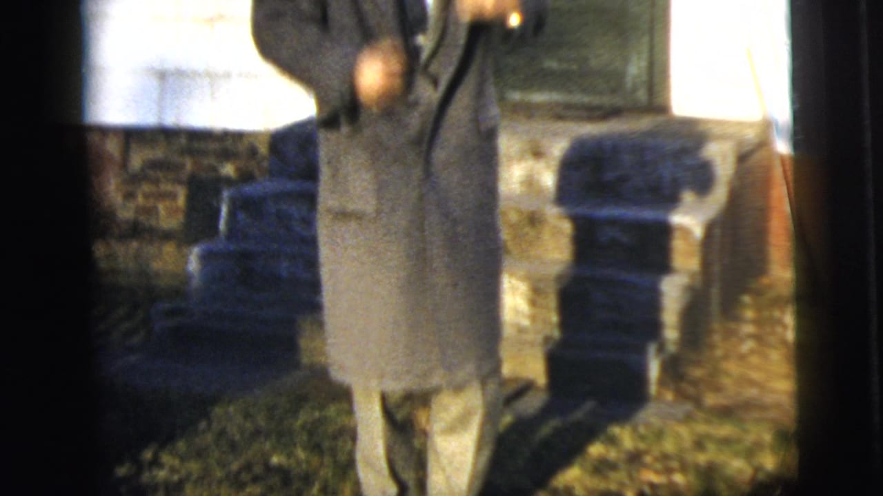 The frame drops lower on the man’s long overcoat and trousers as he stands outside the house in bright daylight.