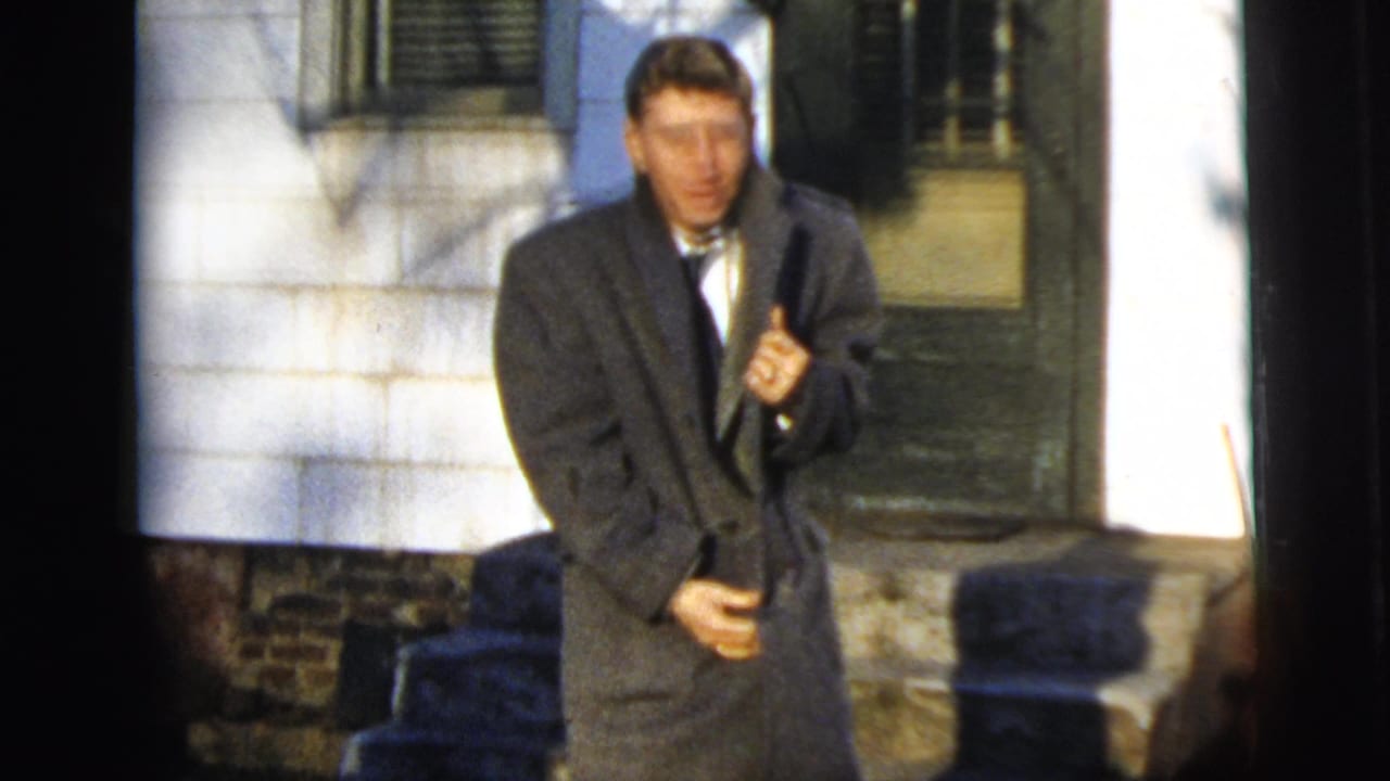 Young man in a gray overcoat standing outside a house doorway.