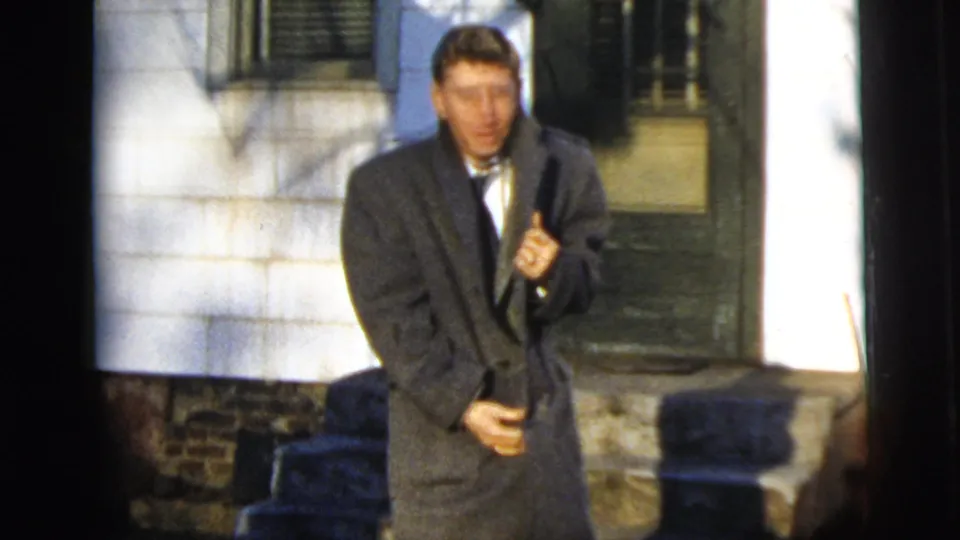 Young man in 1958 New York City home-movie footage standing outside a house in a gray overcoat over formal clothes.