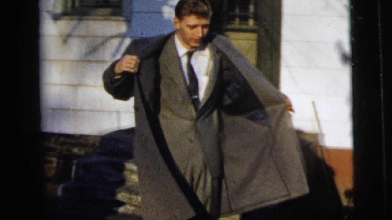 Young man in a gray suit and tie holds his overcoat open while standing outside a house in 1958 New York City footage.