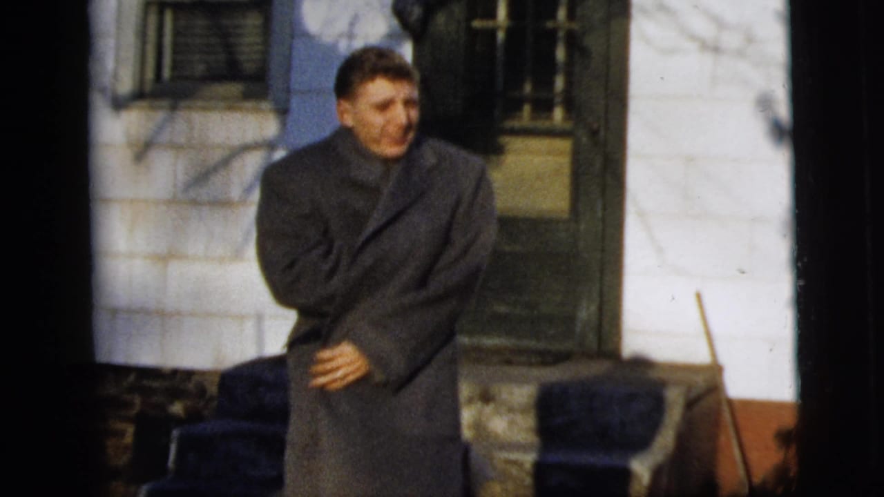 Young man smiles toward the camera while closing or wrapping a gray overcoat outside a New York City house in 1958.