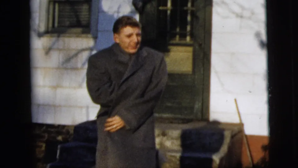 Young man smiles toward the camera while closing or wrapping a gray overcoat outside a New York City house in 1958.