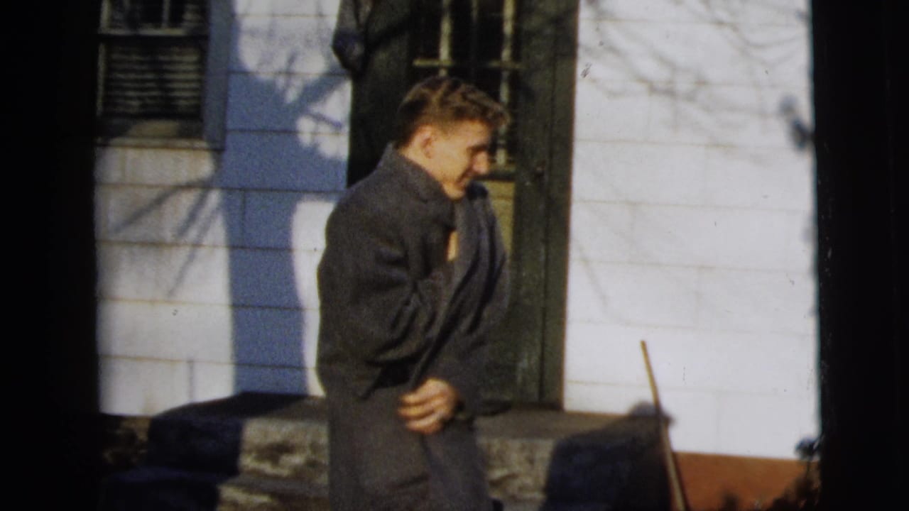 Young man in a dark overcoat stands near a white house exterior and window.