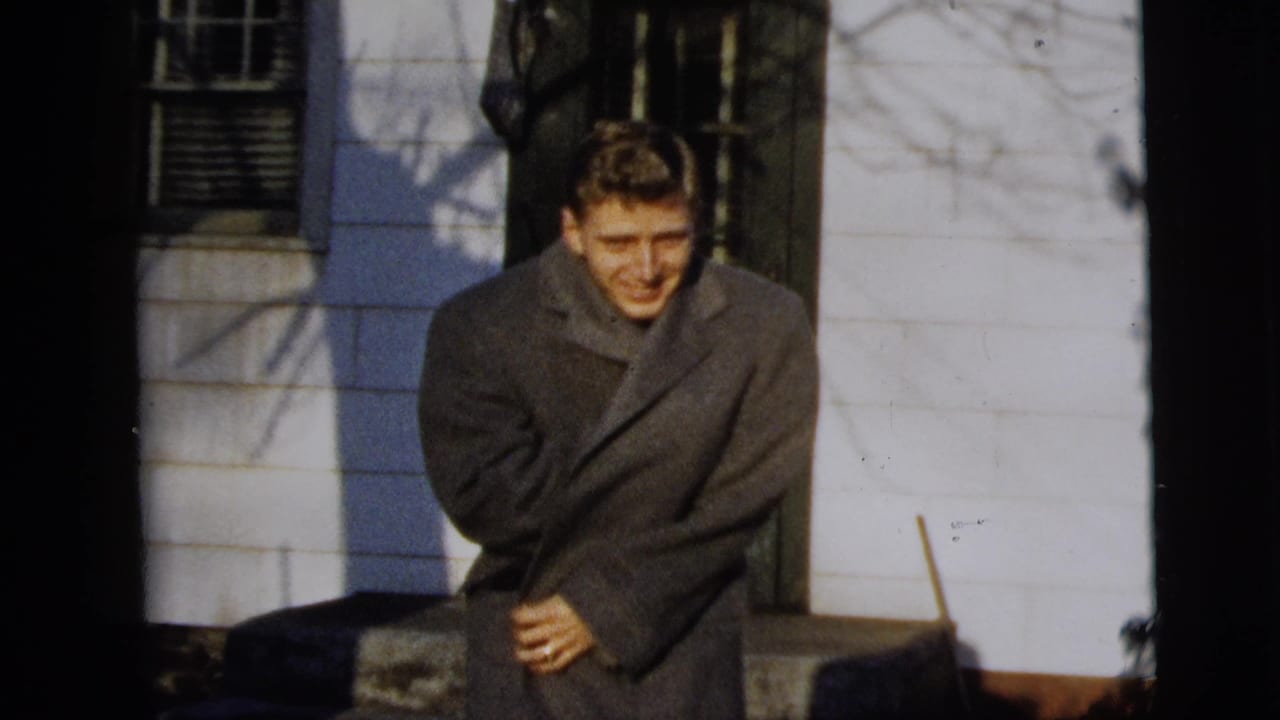 The man smiles and braces in his overcoat outside a white-sided New York City house, with bare branch shadows across the wall.