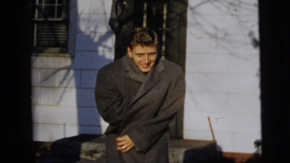 The man smiles and braces in his overcoat outside a white-sided New York City house, with bare branch shadows across the wall.