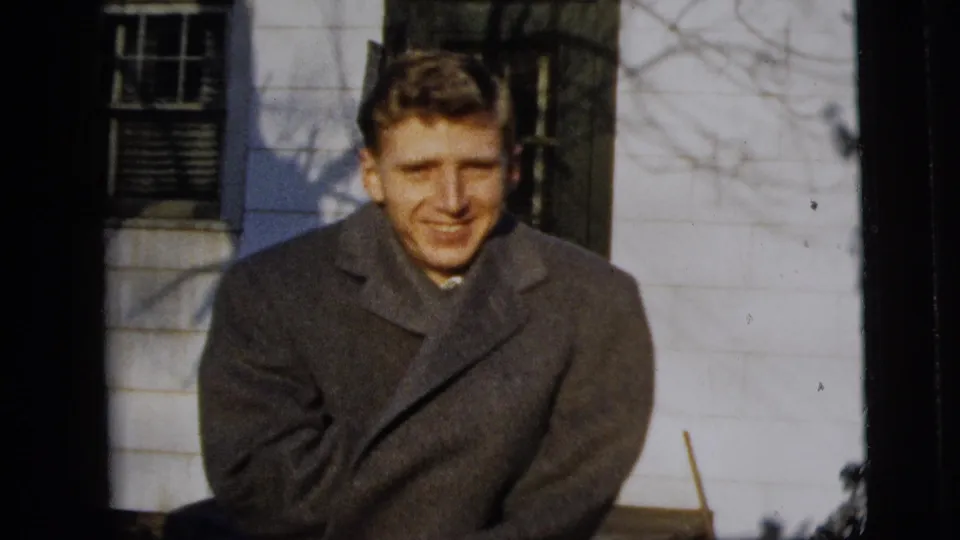 A closer 1958 home-movie frame shows the man facing the camera in a heavy overcoat before a white exterior wall and shuttered window.