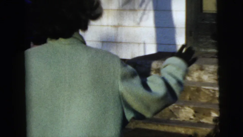 Woman in a pale green coat walks away up exterior steps in a handheld 1958 New York City home-movie frame.
